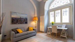 A modern living room with a grey sofa, a desk, a lamp, and a cityscape painting. - GuestHost - Consolata Square Elegant Loft x4 (Torino)