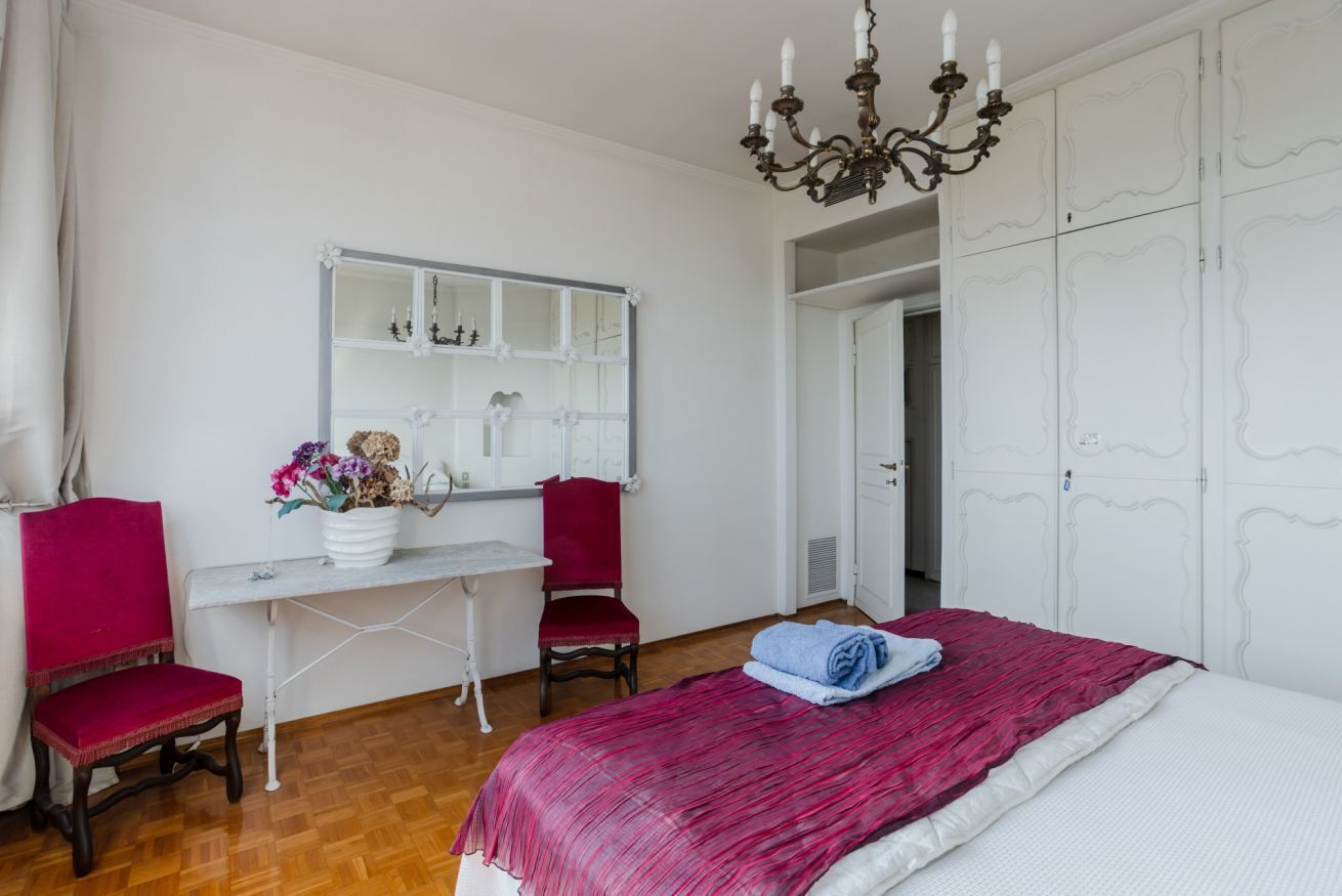 A bedroom with a bed, a desk with a chair, a mirror, and a chandelier.