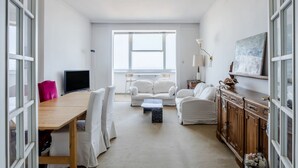 Ett modernt vardagsrum med ett matbord i trä, vita soffor, en tv och ett stort fönster.