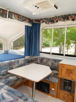 Un intérieur de camping-car compact comprenant un coin cuisine, un espace repas et de grandes fenêtres offrant une vue sur les arbres.