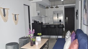 A modern living room with a sofa, ottomans, and a coffee table. There is a kitchen area with a refrigerator and bar stools.
