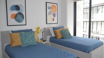 Un hotel moderno con dos camas, cada una con ropa de cama azul y almohadas decorativas. Hay pinturas abstractas en las paredes y un ventanal con vistas a la ciudad.