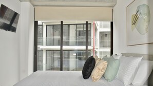 A modern hotel room with a large bed, a view of the building's exterior through floor-to-ceiling windows, and a framed abstract artwork on the wall.