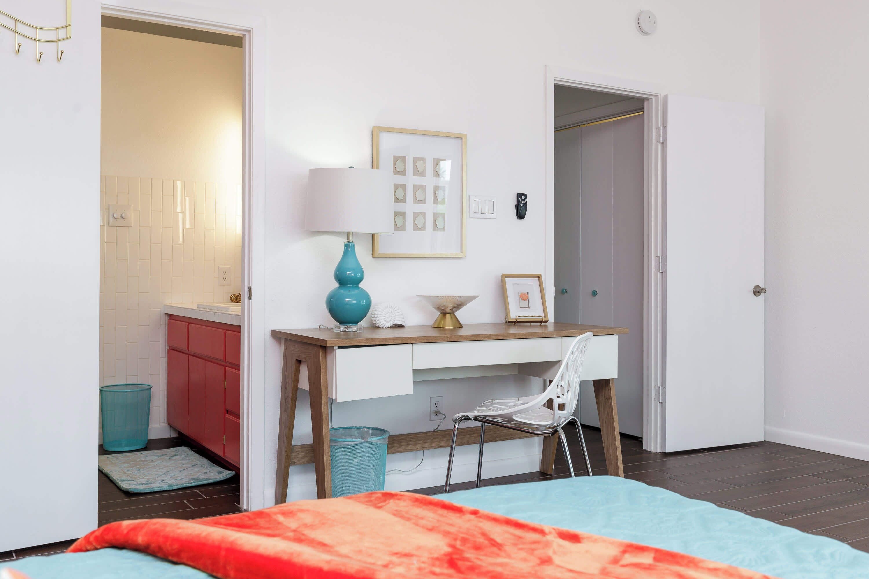 Un dormitorio con cama, escritorio, silla, lámpara, marco de foto y puerta que da al baño.