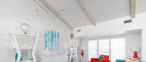 Una sala de estar con pared de madera blanca, una mesa de centro de vidrio, sofás naranja y turquesa, y un estante blanco con objetos decorativos.