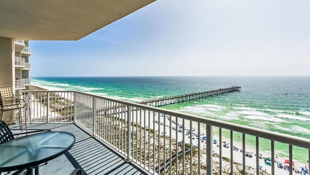 A balcony with a glass table and chairs, overlooking a beach with a pier extending into the ocean.