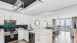 A modern kitchen with white cabinets, a stainless steel stove, and a dishwasher. There is a dining area with a table and chairs, and a balcony with a view.