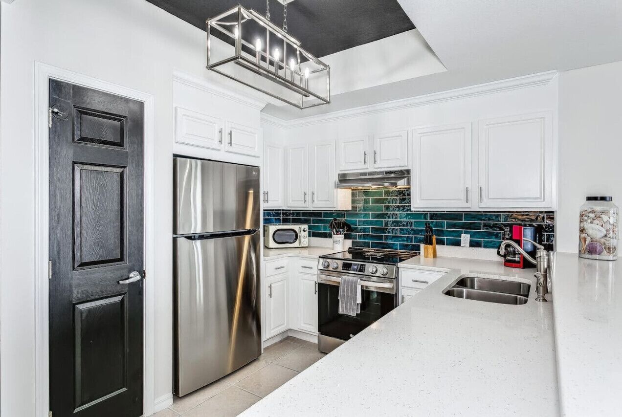 A modern kitchen with white cabinetry, stainless steel appliances, a black door, and a teal backsplash.
