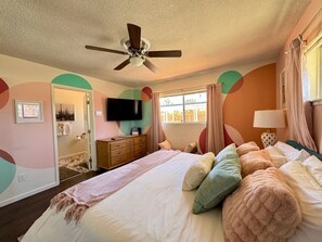 A bedroom with a bed, pillows, a ceiling fan, a dresser, a television, and a window with curtains. - Mid-Century • Pool & Spa • Games • Near JTree Park (Yucca Valley)