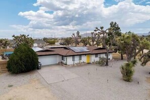 A single-story house with a flat roof, solar panels, and a small yard with desert vegetation. - Mid-Century • Pool & Spa • Games • Near JTree Park (Yucca Valley)