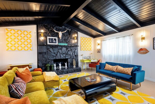 A modern living room with a stone fireplace, a blue sofa, yellow and orange cushions, and geometric wall patterns. - Mid-Century • Pool & Spa • Games • Near JTree Park (Yucca Valley)