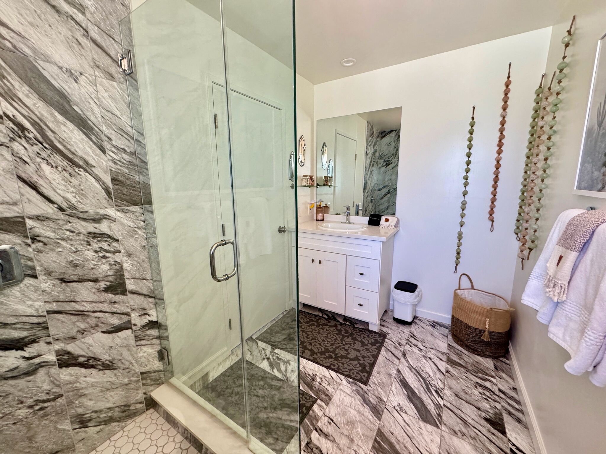 A modern bathroom with a glass shower enclosure, marble flooring, a white vanity with a mirror, and a hanging plant.