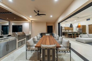 Eine moderne Küche mit integriertem Grill, ein Essbereich mit langem Holztisch und weißen Stühlen sowie eine Wohnbereich mit Sofa und Esstisch.
