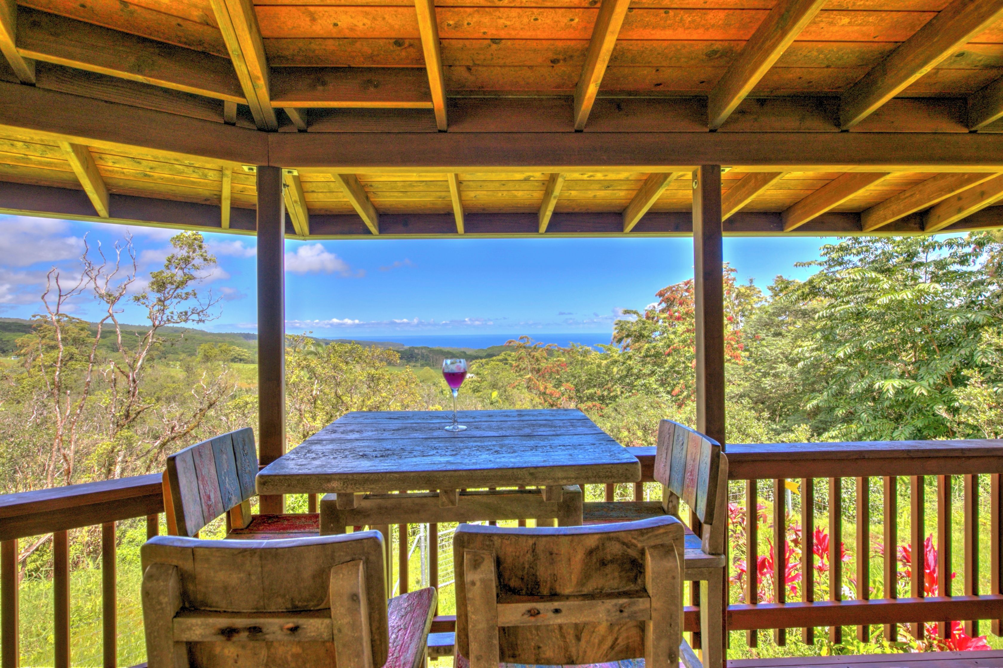 Une table en bois, sur laquelle se trouve un verre de vin rouge, entourée de chaises, sous un toit en bois offrant une vue sur un paysage montagneux.