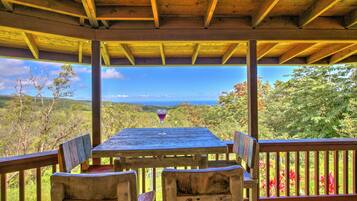 Une table en bois, sur laquelle se trouve un verre de vin rouge, entourée de chaises, sous un toit en bois offrant une vue sur un paysage montagneux.