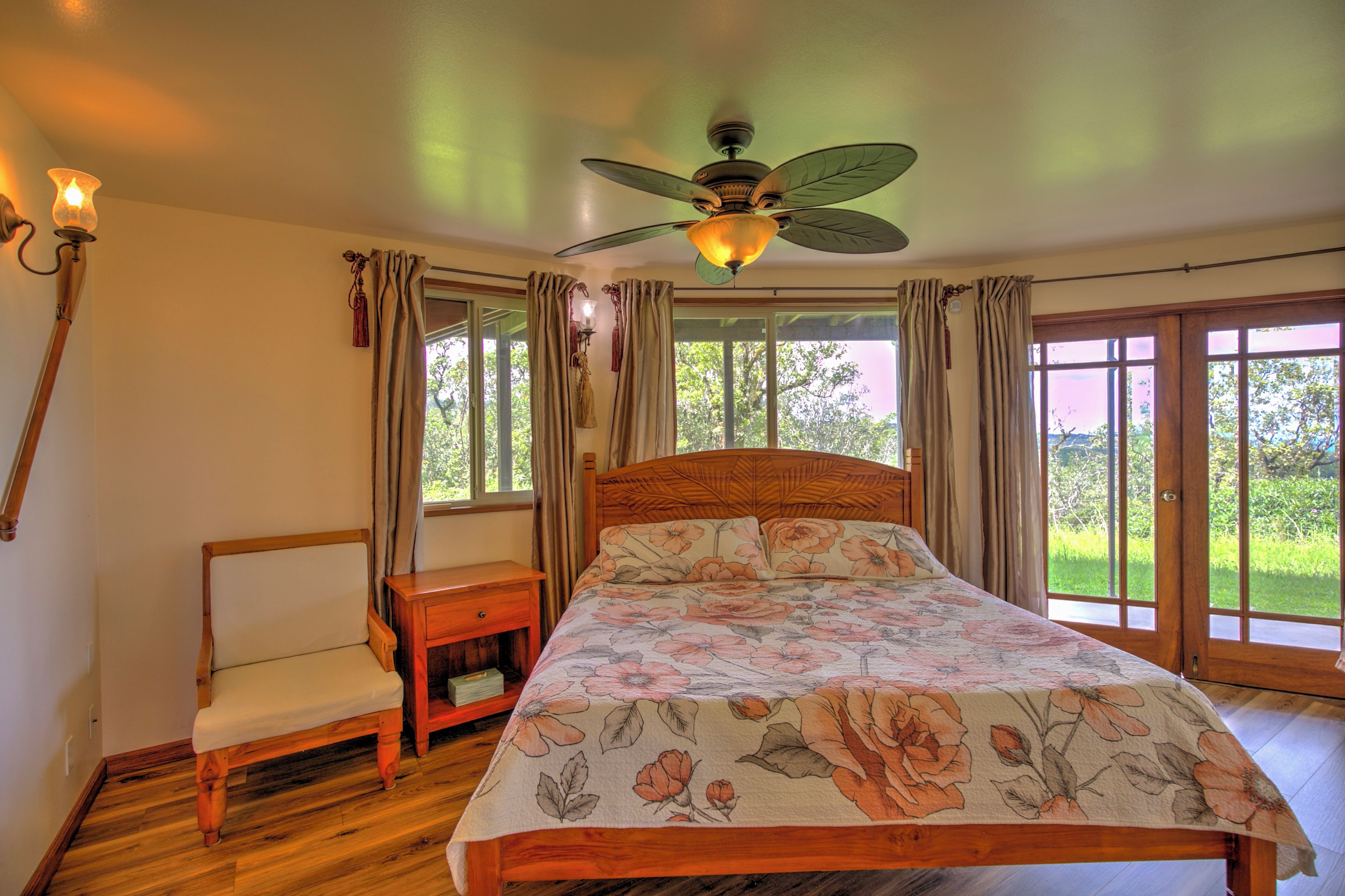 A bedroom with a wooden bed, a nightstand, a chair, a ceiling fan, and large windows with curtains.