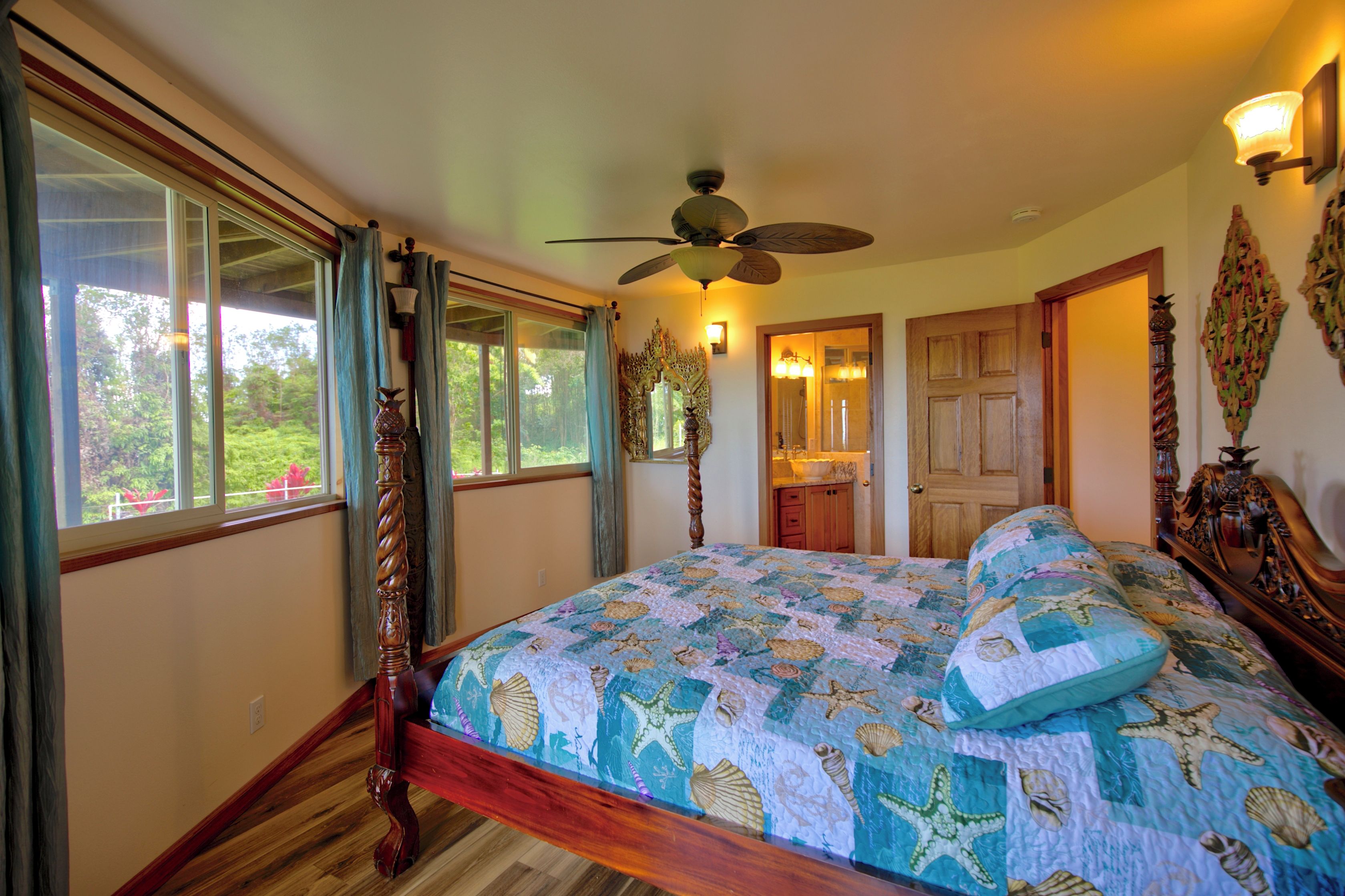 A bedroom with a large bed, a ceiling fan, a view of a kitchen, and a door leading to another room.