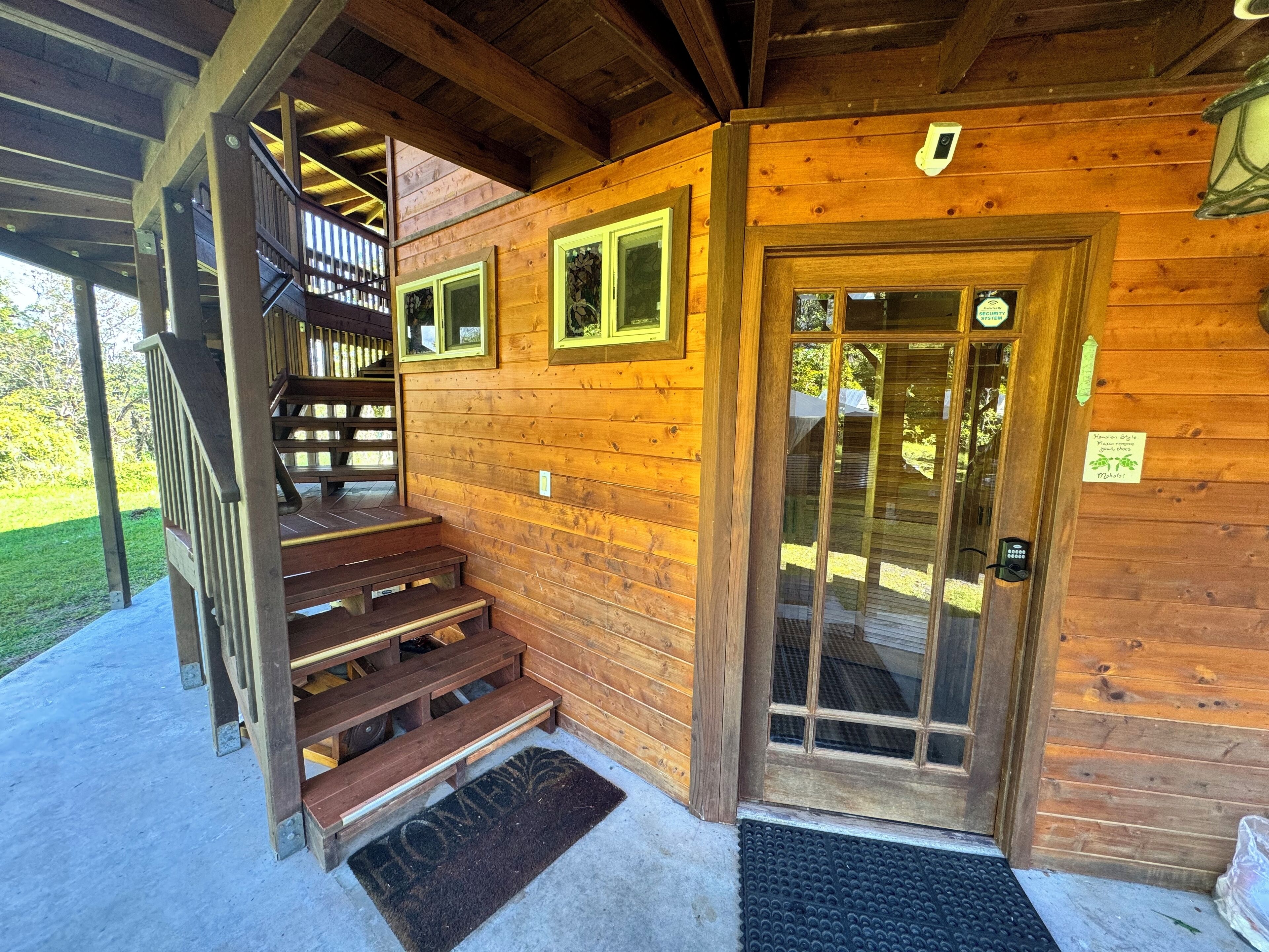 A wooden cabin with a staircase leading to a second level, a door with a security keypad, and a small outdoor area with a bench.
