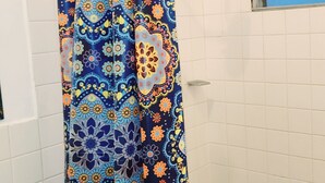 A bathroom with a shower curtain featuring a floral pattern, white tiled walls, a white toilet, and a window with blue blinds. - Hospedaje Casa de Luna (Tegucigalpa)