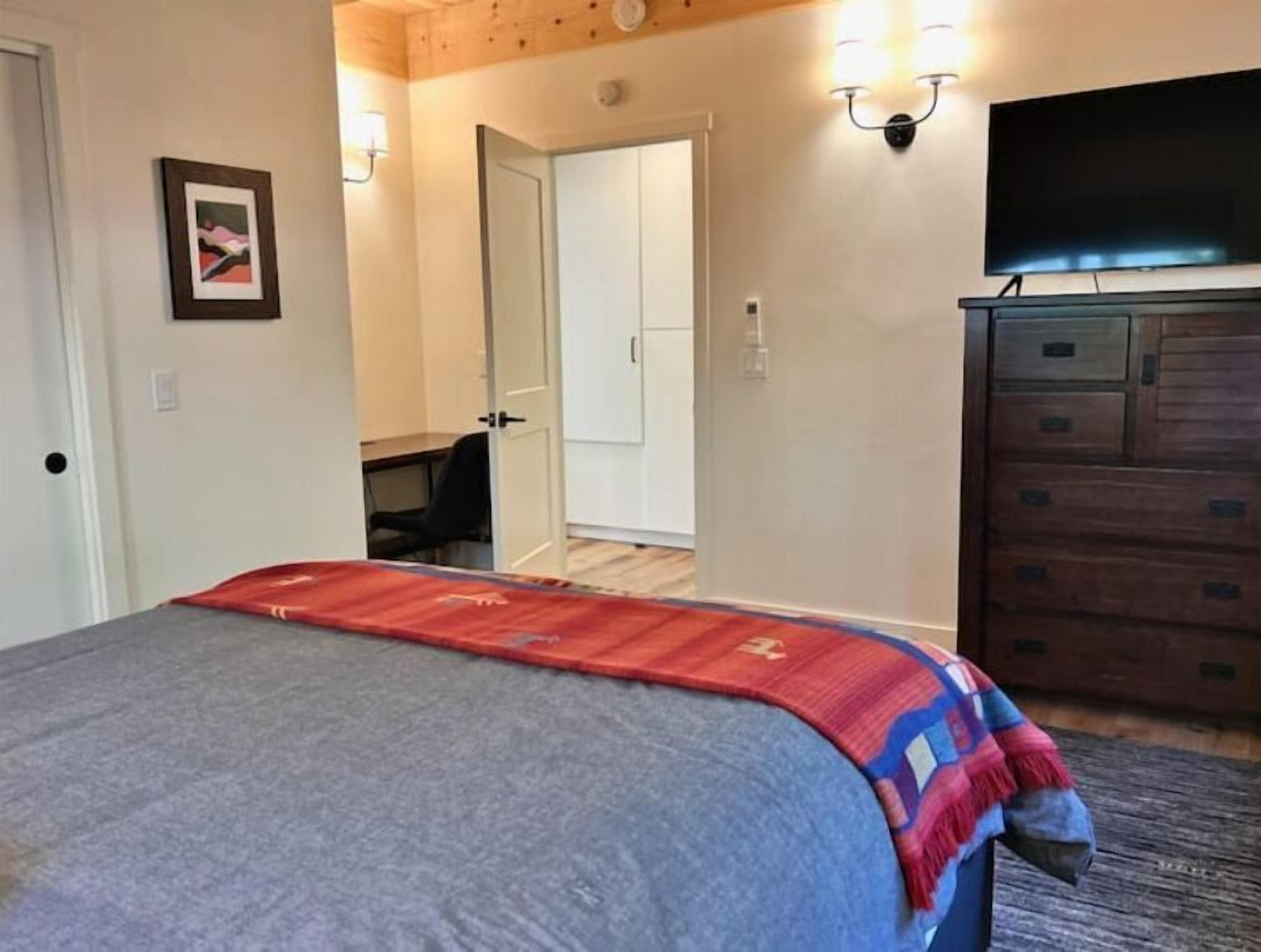 Ein Schlafzimmer mit einem Bett, einer Kommode, einem Fernseher und einer Tür zu einem anderen Raum.