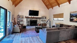 Ein geräumiges Wohnzimmer mit Kamin aus Stein, einem großen Flachbildfernseher und einer gemütlichen Sitzecke mit grauem Sofa und Sessel.