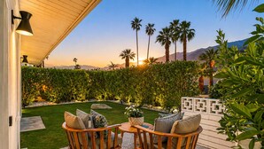 Una veranda con due sedie di legno e un tavolo, circondata da un giardino curato e con vista su palme e montagne.