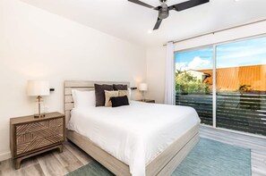 Ein modernes Schlafzimmer mit einem großen Bett, zwei Nachttischen mit Lampen, einem Deckenventilator und einem Balkon mit Blick auf ein Gebäude und Grünflächen.