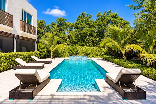 A rectangular swimming pool with clear blue water, surrounded by lounge chairs and a modern building with large windows.