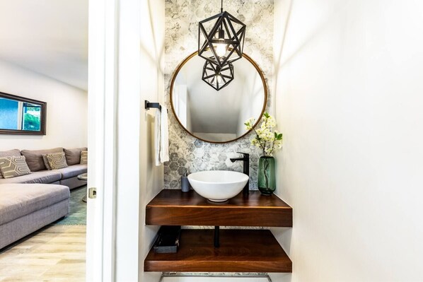Un baño con un espejo redondo, un lavabo blanco tipo recipiente e iluminación colgante geométrica.