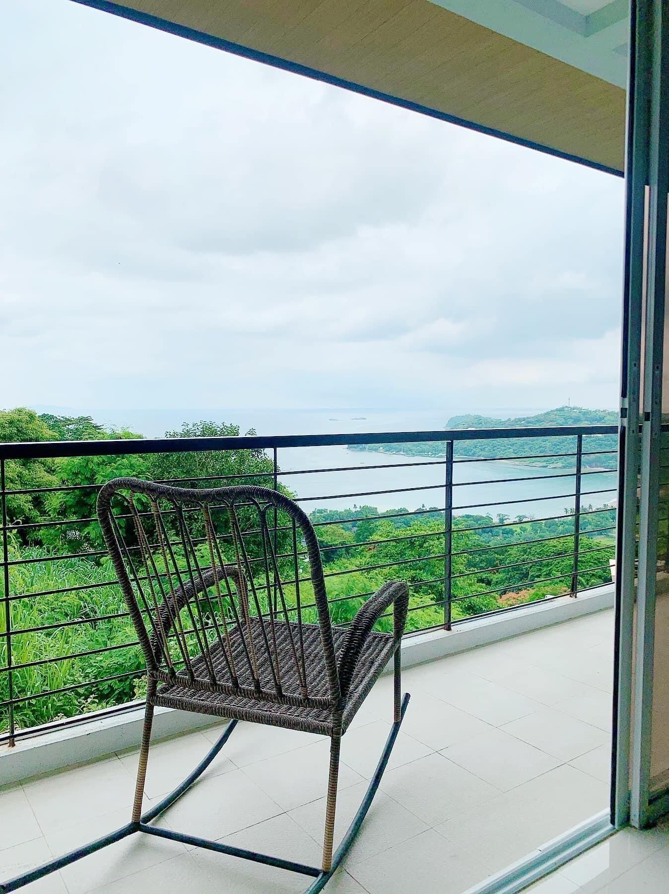 Una silla mecedora de mimbre en un balcón con vista a un lago y follaje.