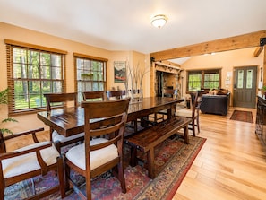 A dining area with a wooden table and chairs, a rug, and a view of the outdoors through large windows.
