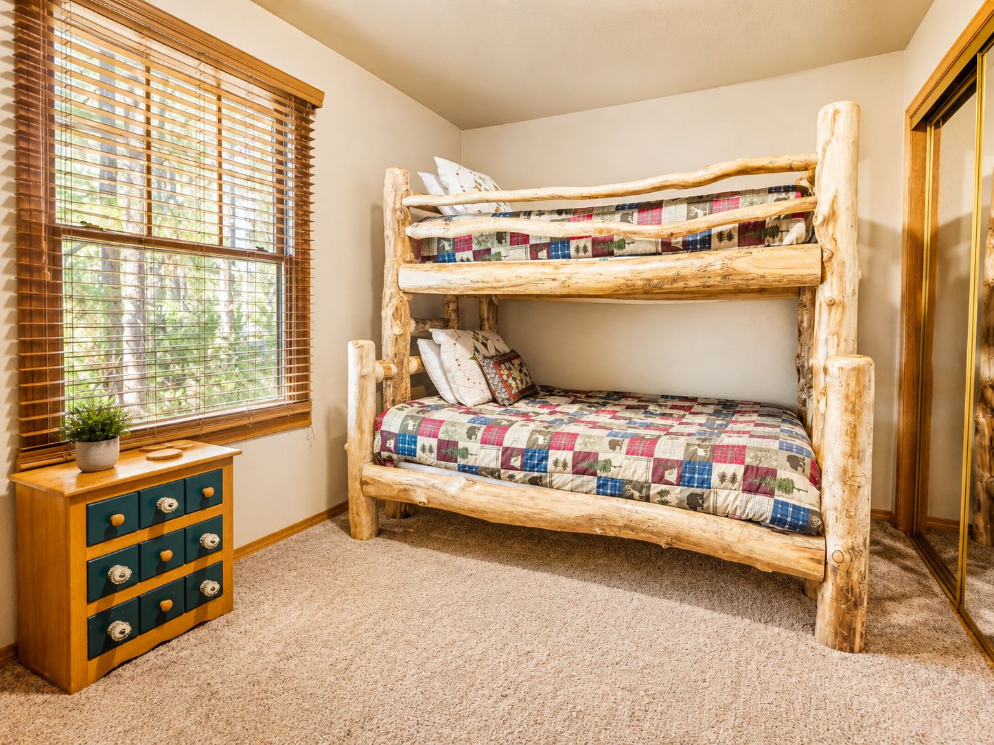 A bunk bed with a patchwork quilt, a wooden dresser with drawers, and a window with blinds.