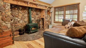 Una sala de estar acogedora con chimenea de piedra, estufa de leña verde, sofá de cuero y una ventana con persianas.