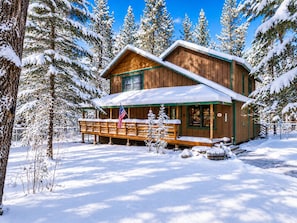 A wooden cabin with a porch and a flag, surrounded by snow-covered trees.