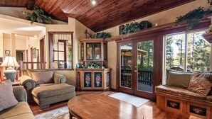 A cozy living room with a wooden coffee table, cushioned seating, a cabinet with floral decorations, and a view through a glass door to the outdoors. - Big Bear Villa - Walk to Lake & Village, Hot Tub, Jet Tub, Outdoor Shower (Big Bear Lake)