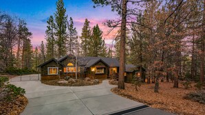 A log cabin with a concrete driveway, surrounded by tall pine trees and a sunset sky. - Big Bear Villa - Walk to Lake & Village, Hot Tub, Jet Tub, Outdoor Shower (Big Bear Lake)