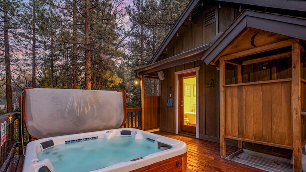A wooden deck with a hot tub, a wooden cabin, and a forested background. - Big Bear Villa - Walk to Lake & Village, Hot Tub, Jet Tub, Outdoor Shower (Big Bear Lake)