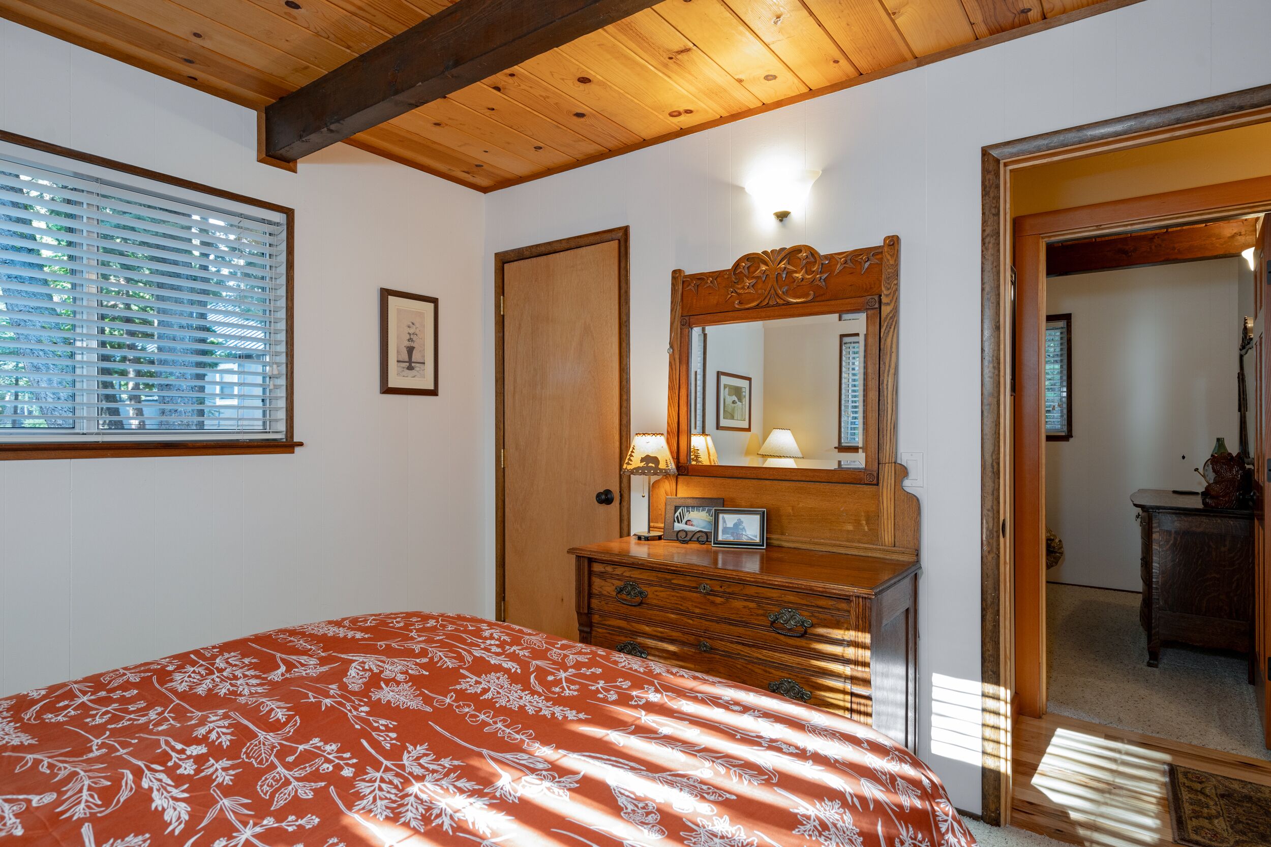 Een slaapkamer met een houten plafond, een bed met een bloemenprint sprei, een kaptafel met spiegel, en een deur die naar een andere kamer leidt.