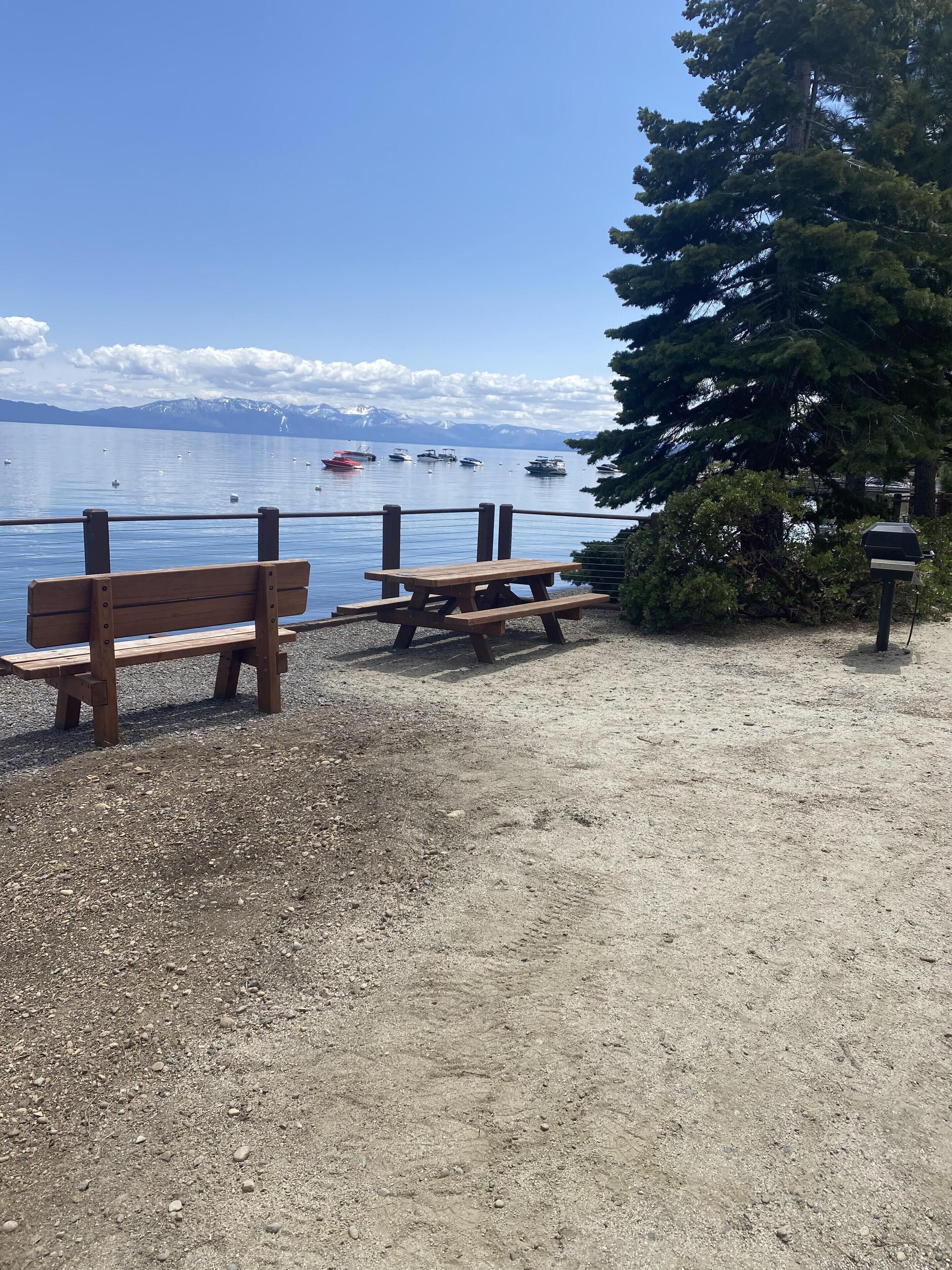 Un espacio de grava con dos mesas de picnic de madera y un banco, con vistas a un lago tranquilo con barcos y montañas nevadas en la distancia.