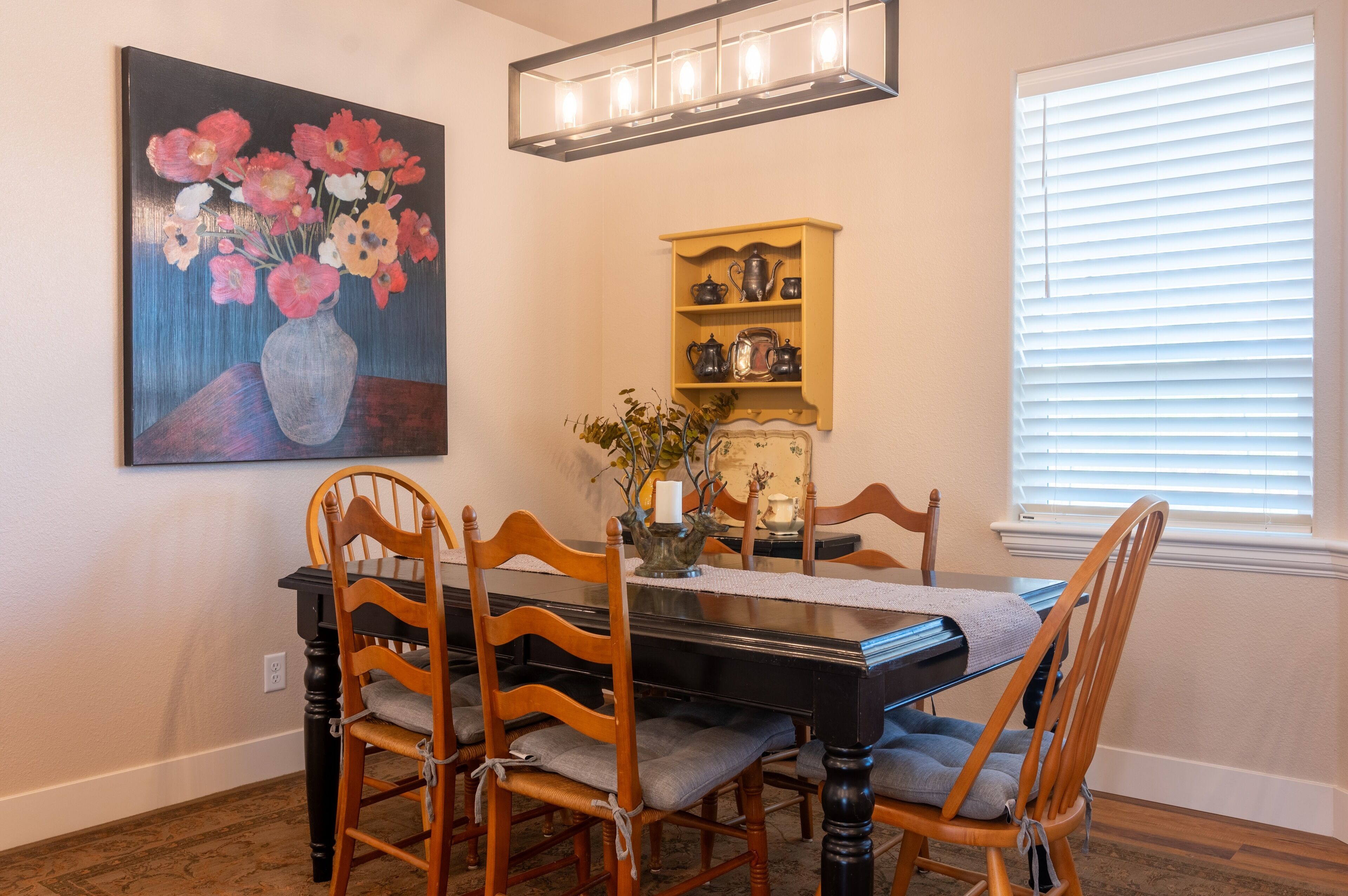 A dining room with a wooden table, chairs, a vase with flowers, a shelf with kitchenware, and a window with blinds.