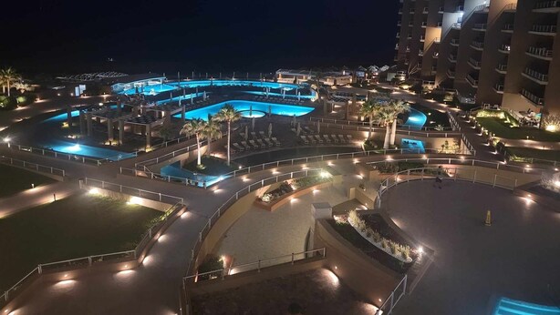 A nighttime view of a hotel with multiple swimming pools, well-lit walkways, and a tall building adjacent to the pool area.