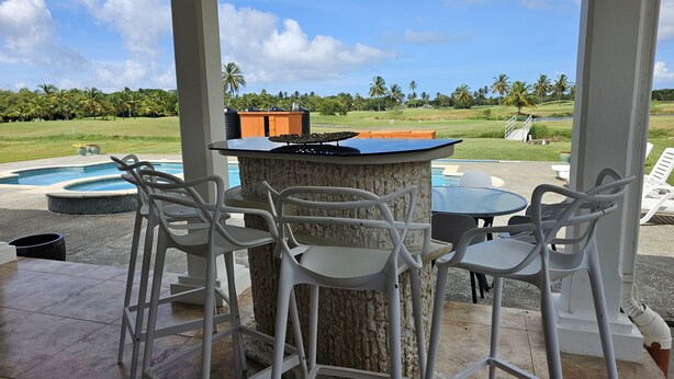 An outdoor bar area with white chairs and a stone table, overlooking a pool and a golf course. - Affordable Ariyana sleeps 10 in Tobago Plantations (Lowlands)