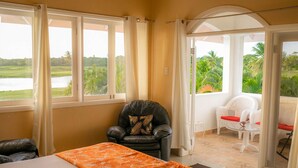A room with a bed, a black leather chair with a decorative pillow, and a view of a golf course and palm trees through large windows. - Affordable Ariyana sleeps 10 in Tobago Plantations (Lowlands)