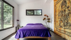 Una habitación con cama, ventana, una pequeña mesa con un florero y un tapiz en la pared.