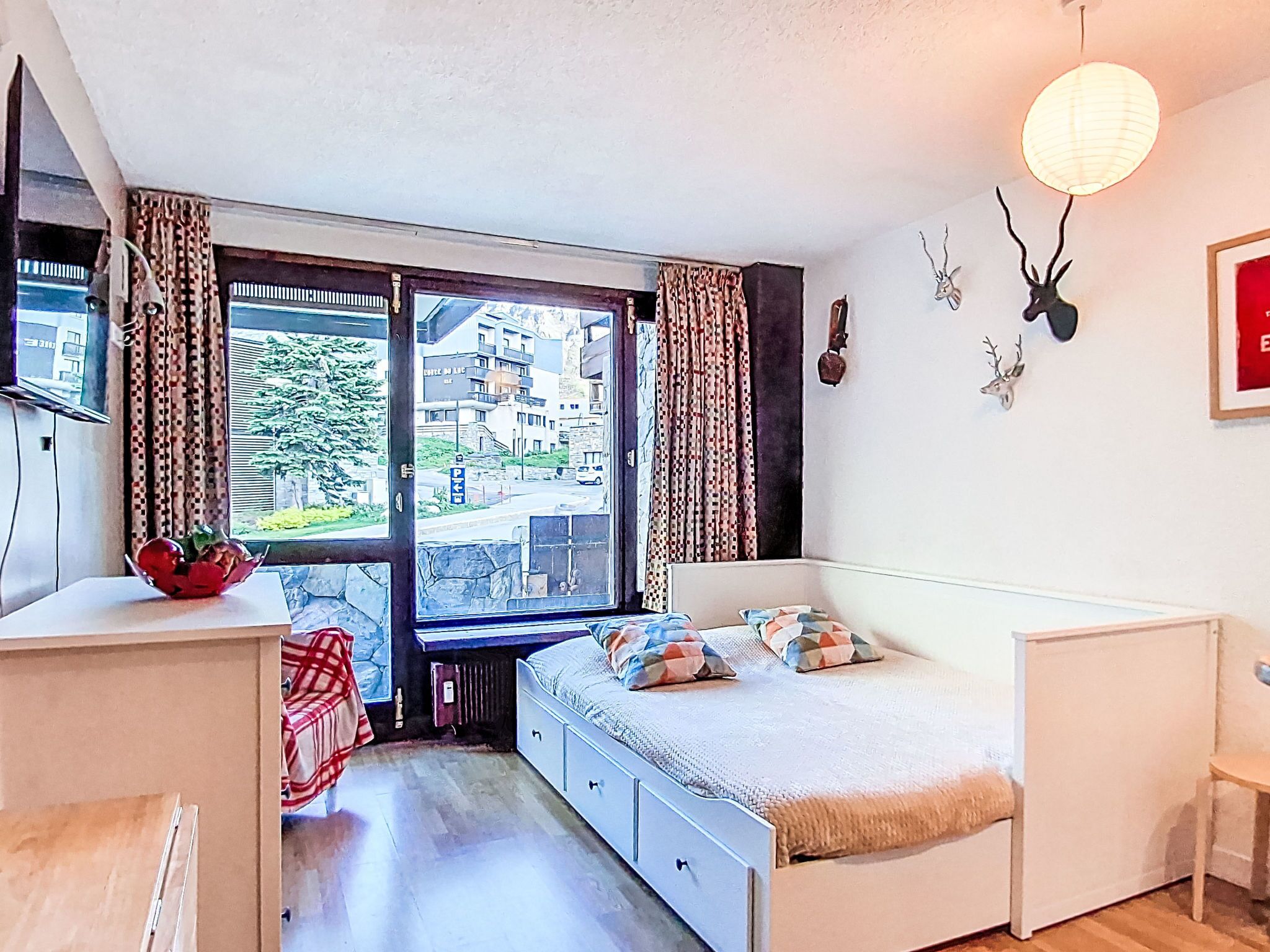 Un dormitorio con cama, escritorio, radiador, ventana con cortinas y vista a edificios.