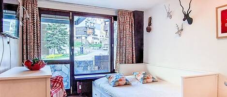 Un dormitorio con cama, escritorio, radiador, ventana con cortinas y vista a edificios.