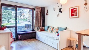 A room with a large window, a white sofa with colorful cushions, a radiator, a small table, and a wall-mounted light fixture. - Les Moutières B1 et B2 (Val Claret) by Interhome (Tignes)