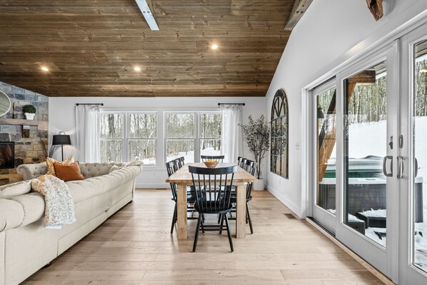 A spacious living area with a wooden ceiling, a beige sofa, a dining table with chairs, and large windows offering a view of the outdoors.