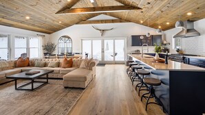 A spacious living room with a large sofa, a coffee table, and a kitchen area with a bar and stools. - ✨ Secluded Luxury w/ Hot Tub, Firepit, Bar + Ultimate Privacy & Views (Ellicottville)