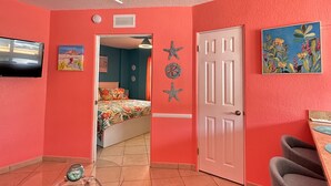 A room with a red wall, a door, a bed with a colorful bedspread, a bicycle, and a chair. - DIRECTLY ON BEACH! JUST STEPS TO THE OCEAN Free Parking Full Kitchen Heated Pool (Pompano Beach)